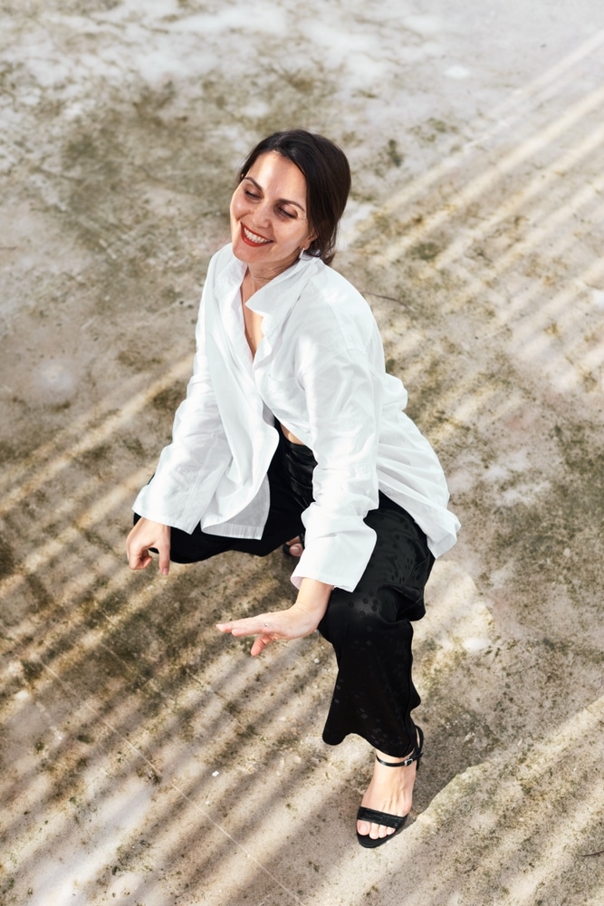 Femme en Chemise Blanche et Pantalon Noir - Mode Chic Femme souriante en chemise blanche et pantalon noir accroupie sur un sol avec des jeux d'ombres.