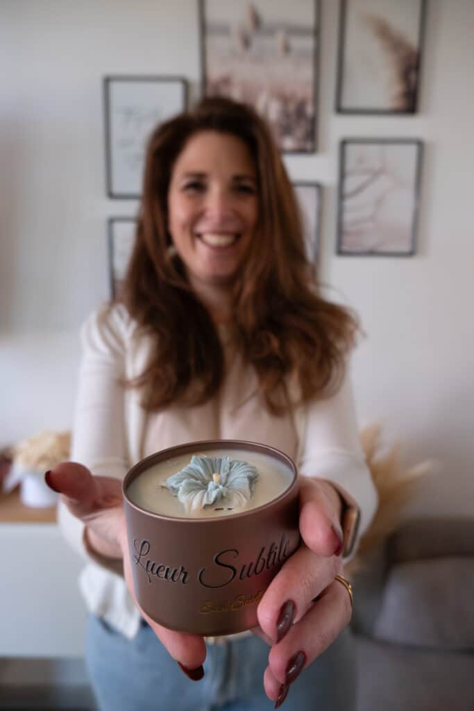 Bougie Artisanale Lueur Subtile : Fleur et Senteur Femme tenant une bougie artisanale Lueur Subtile ornée d'une fleur bleue, ambiance chaleureuse et parfumée.