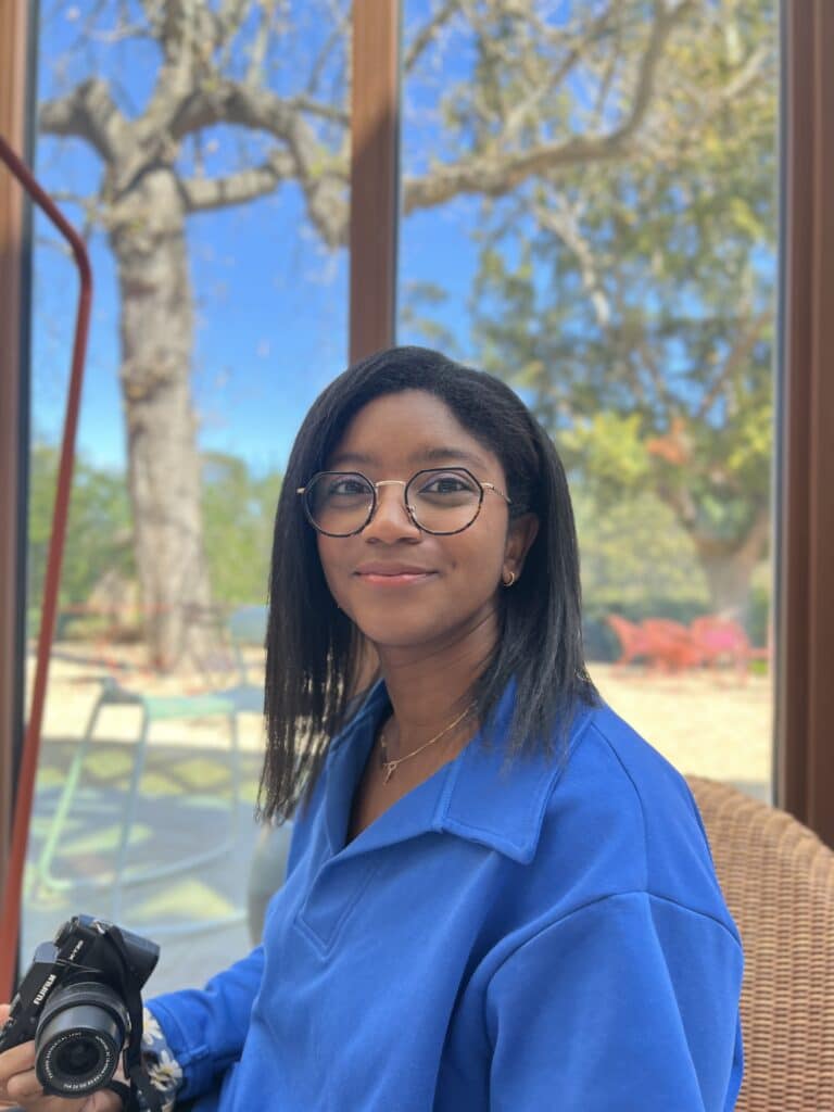 Photographe souriante avec Fujifilm X-T20 et sweat bleu Femme souriante avec lunettes et sweat bleu tenant un appareil photo Fujifilm X-T20 devant une baie vitrée.